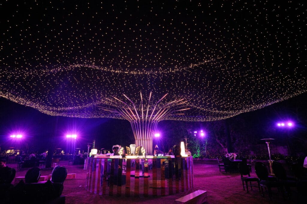 A luxurious outdoor wedding bar with a circular mirrored counter under a massive LED canopy and "tree of lights" installation, designed by a professional Wedding Planner Company in Jaipur.